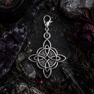 May include: A silver-colored Celtic knot charm with a lobster clasp. The charm is a symmetrical design with interwoven patterns. The background includes crystals, dried flowers, and a lit candle, creating a mystical aesthetic.