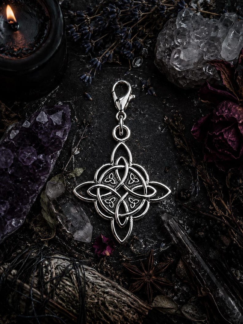 May include: A silver-colored Celtic knot charm with a lobster clasp. The charm is a symmetrical design with interwoven patterns. The background includes crystals, dried flowers, and a lit candle, creating a mystical aesthetic.