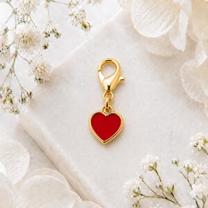 May include: A gold-coloured charm featuring a red heart pendant. The heart is framed in gold and connected to a clasp. The charm is set against a white backdrop with white flowers.