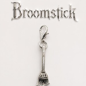 May include: A silver-colored charm in the shape of a broomstick, attached to a clasp. The word "Broomstick" is displayed above the charm in a decorative font. The background is plain white.