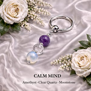 Puede incluir: Un anillo de cuentas cautivas en tono plateado con un colgante vertical de tres cuentas esféricas: una amatista morada, un cuarzo transparente y una piedra lunar blanca lechosa. El texto inferior dice CALM MIND Amethyst · Clear Quartz · Moonstone.