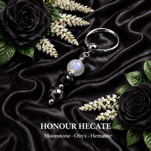 May include: A silver-toned metal ring with a beaded pendant featuring a moonstone, onyx, and hematite. The text "HONOUR HECATE" is displayed below the pendant, with the words "Moonstone • Onyx • Hematite" underneath. The background is black satin with black roses and white flowers.