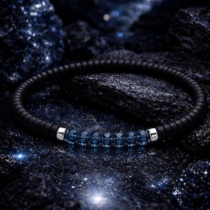 May include: A black bracelet with five faceted blue beads and silver accents. The bracelet is set against a dark, textured background with a starry effect, creating a celestial aesthetic. The bracelet is a piece of jewelry.