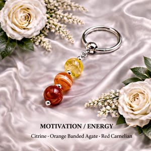 Puede incluir: Un llavero plateado con un colgante de tres cuentas de piedras preciosas: citrino amarillo, ágata bandeada naranja y cornalina roja. El texto "MOTIVATION / ENERGY" y los nombres de las piedras preciosas están en la parte inferior. Rosas blancas y vegetación en el fondo.