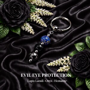 Könnte beinhalten: Ein silberner Schlüsselanhänger mit einem Anhänger aus einer blauen Lapislazuli-Perle, zwei schwarzen Onyx-Perlen und einer Hämatit-Perle. Der Text "EVIL EYE PROTECTION" ist darunter abgebildet. Der Hintergrund ist schwarzer Satin mit schwarzen Rosen und weißen Blumen.