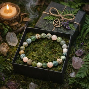Könnte beinhalten: Ein Armband aus grünen, rosa und weißen Perlen liegt in einer schwarzen Schachtel mit grünem Moos. Eine brennende Kerze, Kristalle und eine Geschenkbox mit einem Pentagramm-Anhänger und Farn befinden sich im Hintergrund. Ein natürliches, erdiges Accessoire.