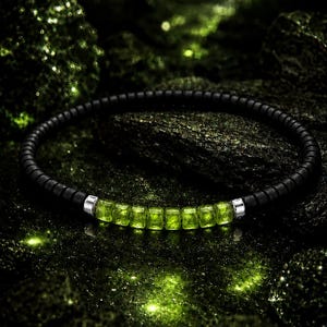 Pode incluir: Uma pulseira de contas pretas com uma fileira de pedras preciosas verdes brilhantes, de corte quadrado. A pulseira tem detalhes metálicos prateados e está colocada sobre um fundo escuro e texturizado com destaques verdes. Uma joia elegante.
