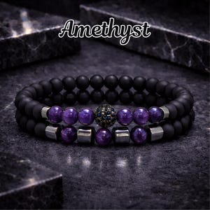 May include: Two beaded bracelets with the word "Amethyst" in white script. The bracelets feature black matte beads, purple amethyst beads, and silver-toned metal accents. One bracelet has a central bead with blue and black stones.