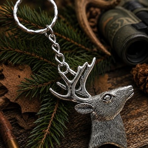 May include: A silver-colored metal keychain featuring a detailed deer head with antlers. The keychain is attached to a silver ring and chain. The deer head is set against a backdrop of pine needles, a wooden surface, and hunting equipment.