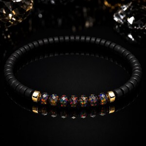 May include: A beaded bracelet featuring a continuous strand of matte black cylindrical beads. The center section includes six faceted, dark iridescent beads with colorful flecks, flanked by two polished gold-tone metal spacers.