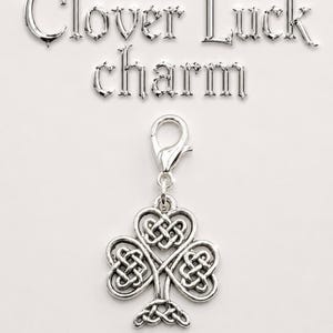 May include: A silver-colored clover charm with a Celtic knot design. The charm is attached to a clasp and is displayed against a white background. The words "Clover Luck charm" are written above the charm.