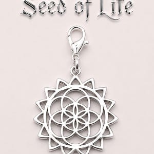 May include: A silver charm pendant with a sunburst design and a central geometric pattern. The pendant is attached to a clasp. The words "Seed of Life" are displayed above the charm in a stylized font.