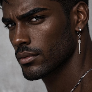 May include: A close-up portrait of a person with dark skin, wearing a silver arrow earring and a silver chain necklace. The earring is a small hoop with a dangling arrow pendant. The person has a groomed beard and moustache.