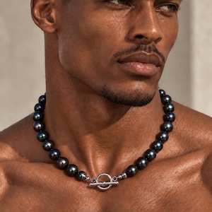 May include: A necklace featuring round, dark, iridescent black pearls measuring 8 mm in diameter. The strand is secured with a polished silver-toned metal toggle clasp consisting of a circular ring and a horizontal bar. The text reads BLACK PEARL 8 MM BEADS.