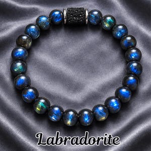 Peut inclure: Un bracelet de perles en labradorite avec des sphères bleues et vertes irisées. Le bracelet comporte une perle cylindrique noire et des accents argentés. Le texte "Labradorite" est affiché en bas de l'image.