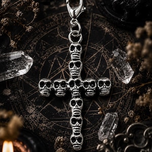 May include: A silver-colored cross-shaped charm featuring skull designs. The charm has a lobster clasp for attachment and is surrounded by crystals and candles on a dark, patterned surface. The gothic-inspired accessory is suitable for jewelry or bag charms.