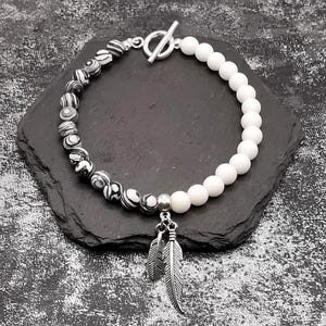 Könnte beinhalten: Armband mit einer Mischung aus schwarz-weiß gemusterten Perlen und weißen Perlen. Es hat einen silbernen Knebelverschluss und einen Anhänger mit zwei Federanhängern. Das Armband wird auf einer dunkelgrauen Oberfläche präsentiert.