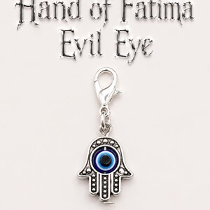 May include: A silver-toned charm with a Hand of Fatima design and an evil eye symbol. The charm has a lobster clasp and features a blue and black eye in the center of the hand. The words "Hand of Fatima Evil Eye" are above the charm.