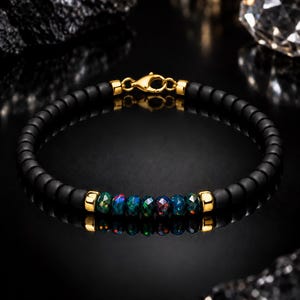 May include: A beaded bracelet featuring matte black round beads and a central row of faceted dark opal beads with iridescent flashes of color. The bracelet is accented with polished gold-tone metal spacers and finished with a gold-tone lobster claw clasp.