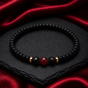 Puede incluir: Una pulsera de cuentas con cuentas esféricas de color negro mate, dos cuentas negras facetadas y una cuenta roja central facetada. La pulsera incluye pequeños espaciadores cilíndricos de tono dorado entre las cuentas facetadas.