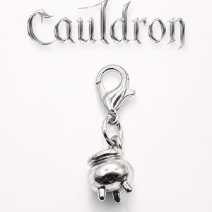 May include: A silver-colored charm featuring a small cauldron with three legs, attached to a clasp. The word "Cauldron" is displayed above the charm in a stylized, metallic font against a white background.