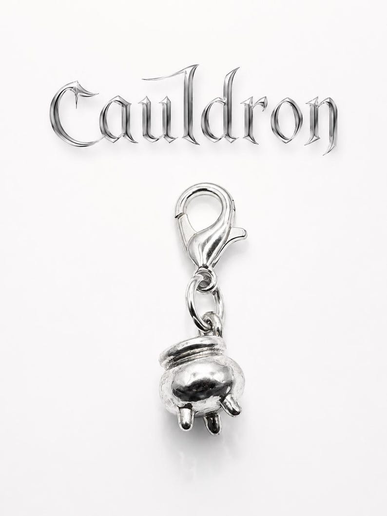 May include: A silver-colored charm featuring a small cauldron with three legs, attached to a clasp. The word "Cauldron" is displayed above the charm in a stylized, metallic font against a white background.