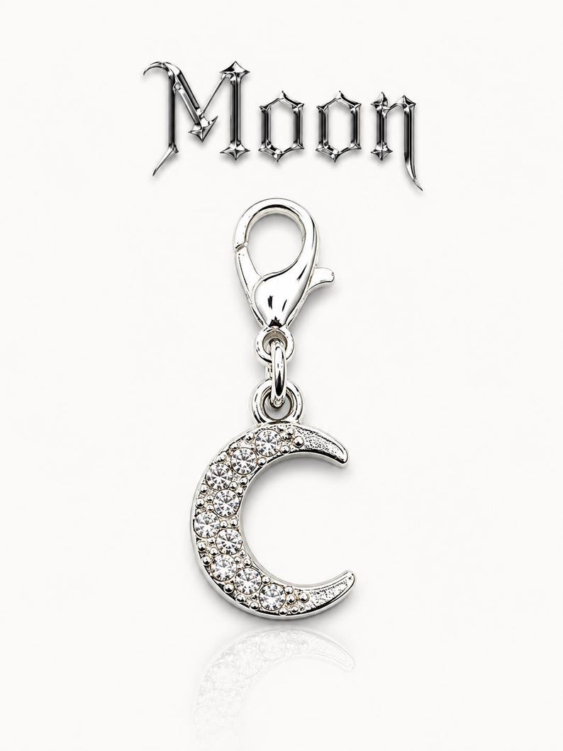 May include: A silver-colored charm in the shape of a crescent moon, encrusted with clear crystals. The charm is attached to a clasp. The word "Moon" is displayed above the charm in a decorative font.
