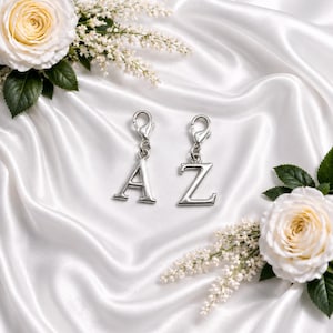 Könnte beinhalten: Zwei silberne Buchstabenanhänger, ein "A" und ein "Z", mit Karabinerverschlüssen, liegen auf einer weißen Satin-Oberfläche. Weiße Rosen und zarte weiße Blüten mit grünen Blättern umgeben die Anhänger.