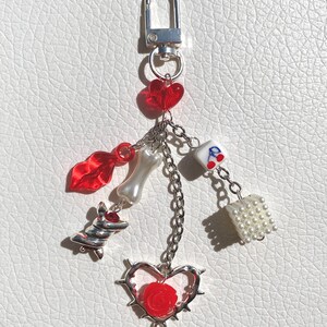 May include: A silver keychain with a variety of charms, including a red heart, a red lip, a white bone, a white dice charm, a white bead charm, a red rose in a silver heart with thorns, and a silver swirl charm.