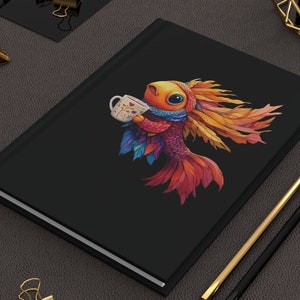 May include: A black hardcover notebook featuring a colorful illustration of a fish with orange, red, and blue scales holding a small white mug with a floral pattern. The notebook is positioned next to two pencils and several gold binder clips.