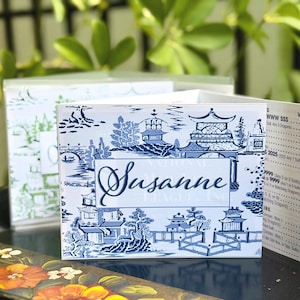 May include: A folded card featuring a blue and white chinoiserie pattern with pagodas, bridges, and trees. The front displays the name Susanne in a dark blue script font within a rectangular border. The side panel shows a printed list of game values.