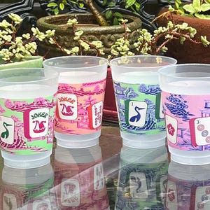 May include: Four clear plastic cups with colorful designs. Each cup features a different playing card design with the word "Joker" and other card symbols. The cups are filled with a clear liquid and are set on a reflective surface.