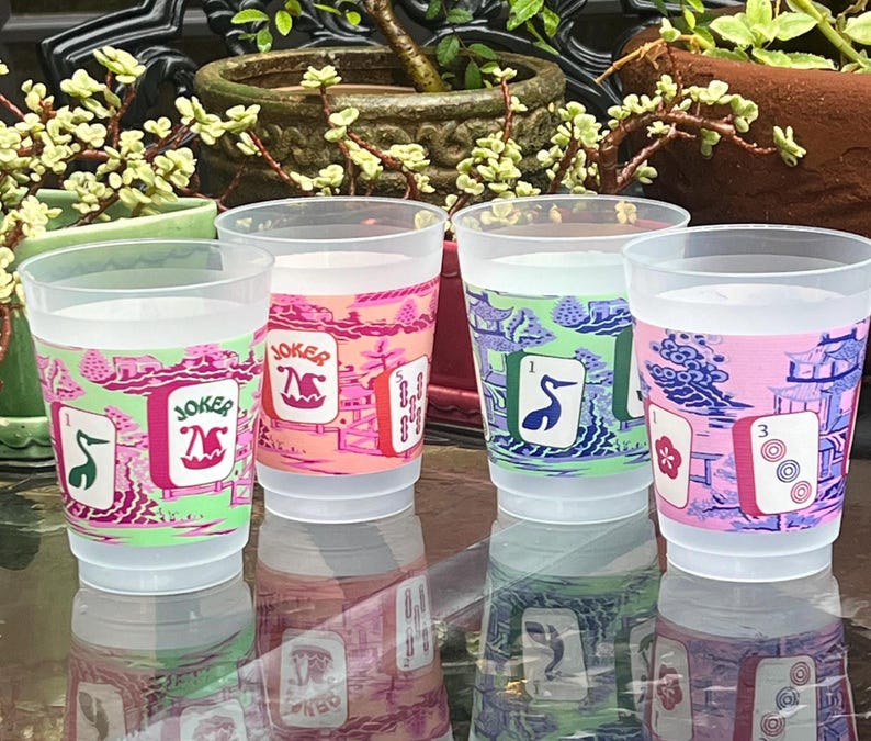 May include: Four clear plastic cups with colorful designs. Each cup features a different playing card design with the word "Joker" and other card symbols. The cups are filled with a clear liquid and are set on a reflective surface.