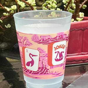 May include: A clear plastic cup with a pink and peach-colored band featuring a stylized Asian landscape design. The band includes two playing card images: a pink crane and a "JOKER" card. The cup is filled with a clear liquid.