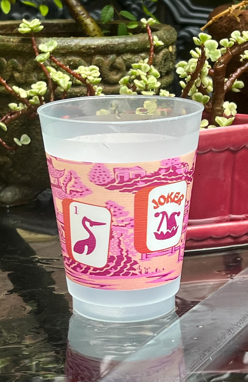 May include: A clear plastic cup with a pink and peach-colored band featuring a stylized Asian landscape design. The band includes two playing card images: a pink crane and a "JOKER" card. The cup is filled with a clear liquid.