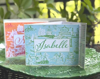 May include: Two personalized cards with the name "Isabelle" on the front. One card is light blue with a green Asian-inspired design, and the other is orange with a white design. The cards are on a green leaf-shaped plate.