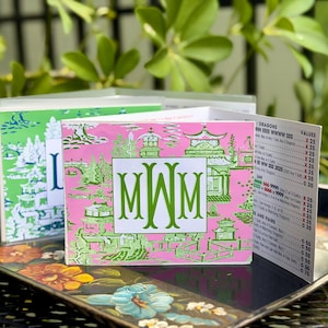 May include: Two decorative cards with a pink and green Asian-inspired design. The pink card features a green monogram and pagoda illustrations. The green card has a similar design. A price list is visible. The cards are on a floral tray.