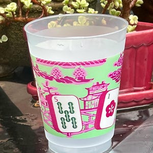 May include: A clear plastic cup filled with liquid, featuring a decorative design in pink and green. The design includes stylized Asian-inspired imagery, including pagodas and playing card symbols. The cup has the numbers "1" and "5" on the design.
