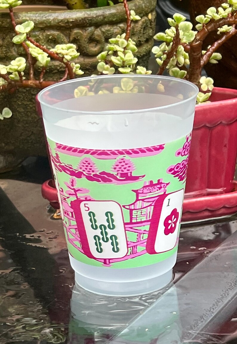 May include: A clear plastic cup filled with liquid, featuring a decorative design in pink and green. The design includes stylized Asian-inspired imagery, including pagodas and playing card symbols. The cup has the numbers "1" and "5" on the design.