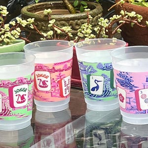 May include: Four clear plastic cups with colorful designs. Each cup features a different design, including the word "Joker" and illustrations of playing cards. The cups are decorated with pink, green, and blue hues.