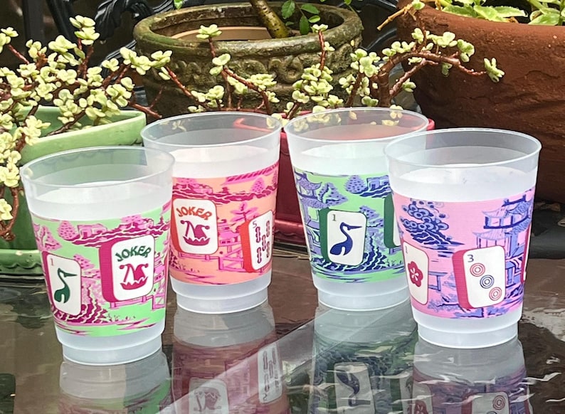 May include: Four clear plastic cups with colorful designs. Each cup features a different design, including the word "Joker" and illustrations of playing cards. The cups are decorated with pink, green, and blue hues.