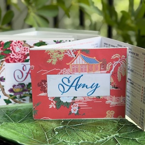 May include: Two decorative place cards with floral and Asian-inspired designs. One card features a red background with an Asian pagoda and the name "Amy" in blue script. The other card has a floral pattern with pink and purple flowers. Both cards are open.