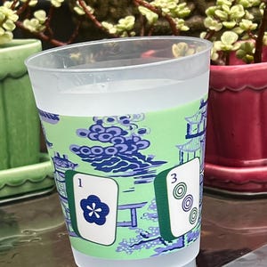 May include: A clear plastic cup with a green and blue patterned design. The design includes a pagoda and Mahjong tiles. One tile shows a flower and the number 1, and another shows the number 3 with three circles.