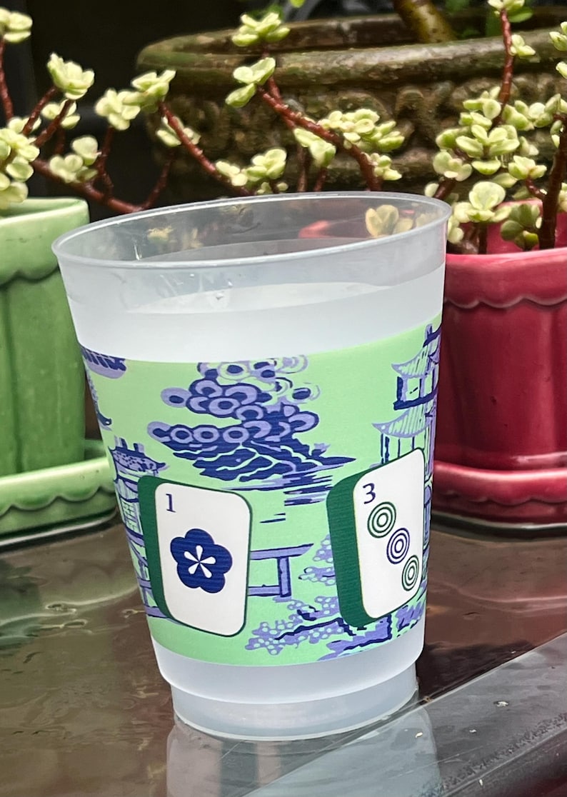 May include: A clear plastic cup with a green and blue patterned design. The design includes a pagoda and Mahjong tiles. One tile shows a flower and the number 1, and another shows the number 3 with three circles.