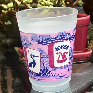 May include: A clear plastic cup with a pink and blue decorative band. The band features a landscape design and the word "JOKER" with a jester hat. One side shows a card with the number 1 and a blue bird.