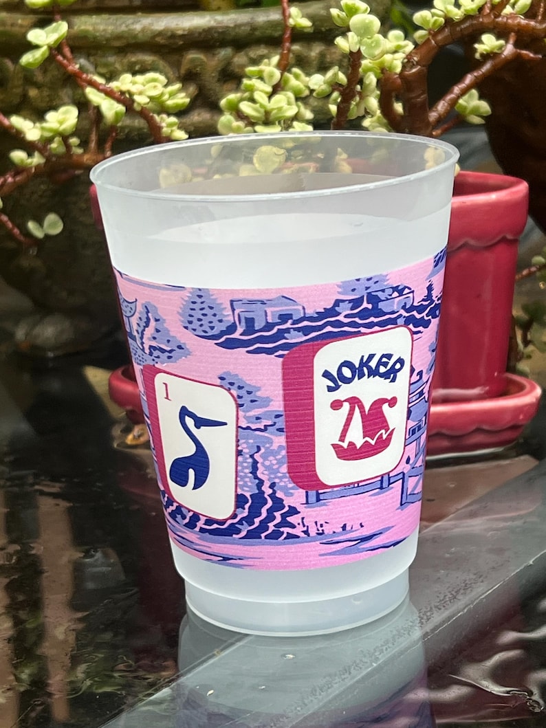 May include: A clear plastic cup with a pink and blue decorative band. The band features a landscape design and the word "JOKER" with a jester hat. One side shows a card with the number 1 and a blue bird.