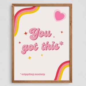 Op de afbeelding: Een ingelijst print met een roze achtergrond en de tekst "You got this*" in roze met een roze hart en regenboogstrepen op de achtergrond. De tekst "*crippling anxiety" staat onderaan de print.