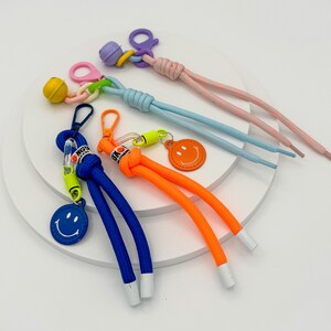 May include: Four colorful shoelace charms with smiley face designs. The charms are attached to shoelaces in blue, orange, pink, and light blue. The shoelaces are tied together and arranged on a white surface.