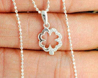 Puede incluir: Un collar de cadena de plata con un colgante de trébol de cuatro hojas. El trébol está bordeado de pequeñas piedras transparentes.