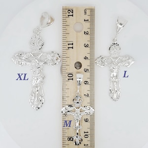 May include: Three silver filigree cross pendants of different sizes, labelled "XL", "M", and "L", are shown next to a ruler. The ruler is marked in inches and centimetres.